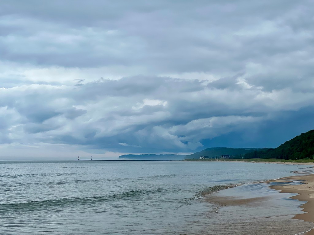 A serene view of a beach with gentle waves lapping at the shore, under a cloudy sky that hints at an impending storm. Lighthouses are visible in the distance along the coastline.