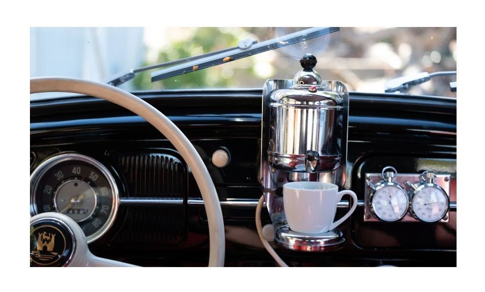 A vintage coffee maker mounted in the dashboard of a classic car, with a white coffee cup positioned underneath it.