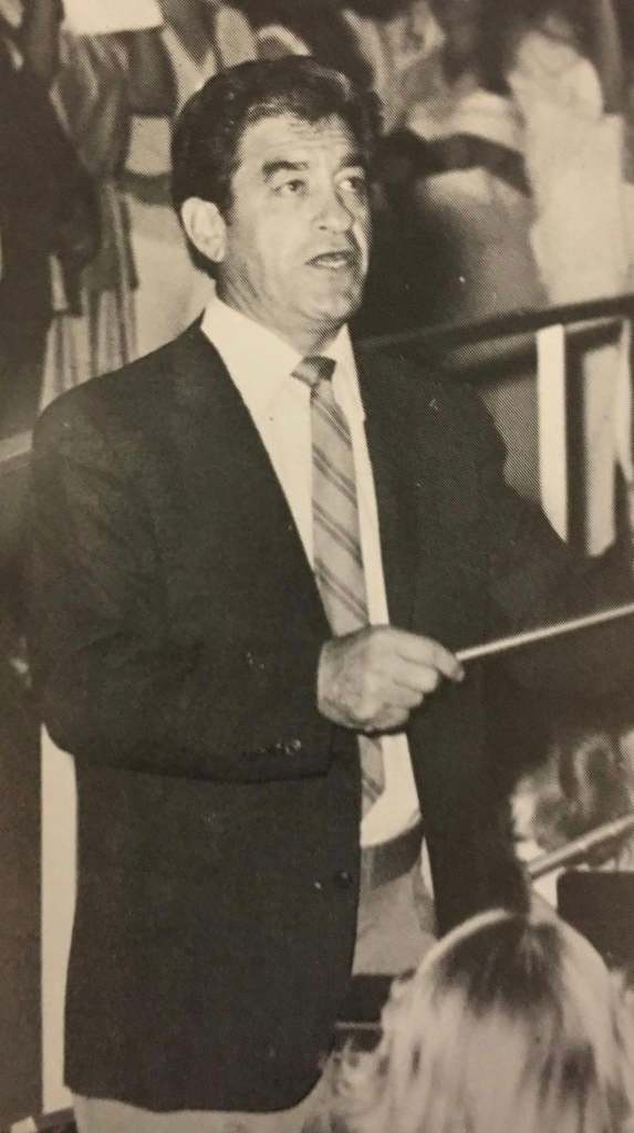 A man in a suit and tie addressing an audience, holding a conducting baton.