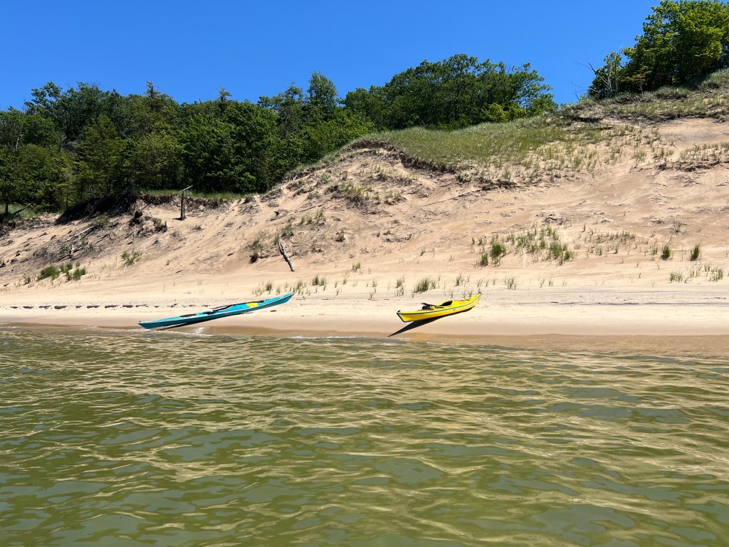 Two kayaks, one blue and one yellow, resting on a sandy beach with a lush green hillside in the background under a clear blue sky.