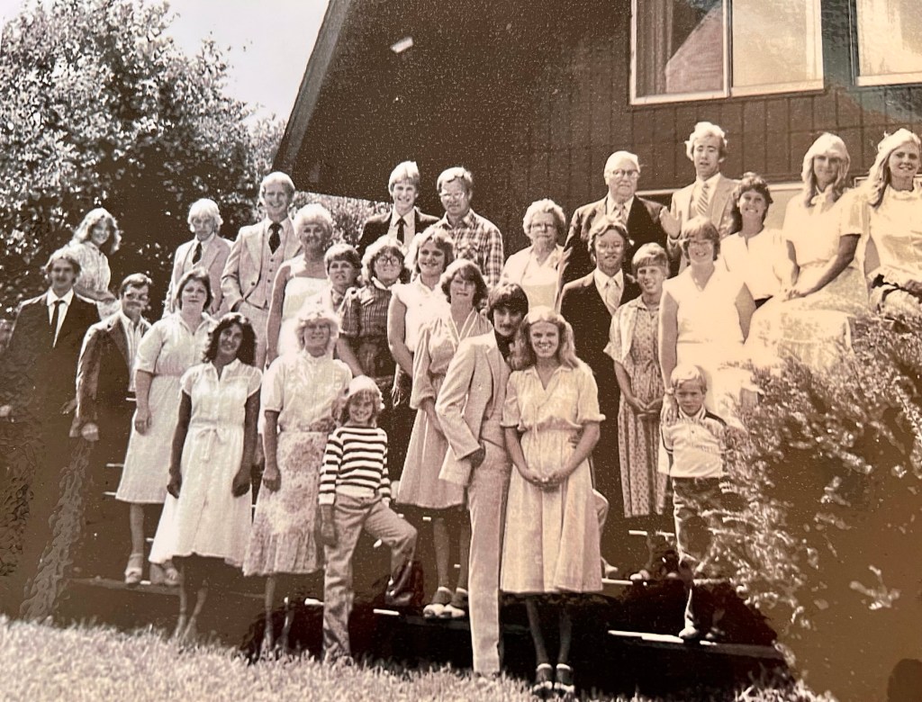 A large group of people gathered outdoors in front of a wooden building, posing for a photo. The group includes men, women, and children, dressed in a variety of formal and casual clothing, with some wearing white dresses. The setting features greenery and a sunny atmosphere.