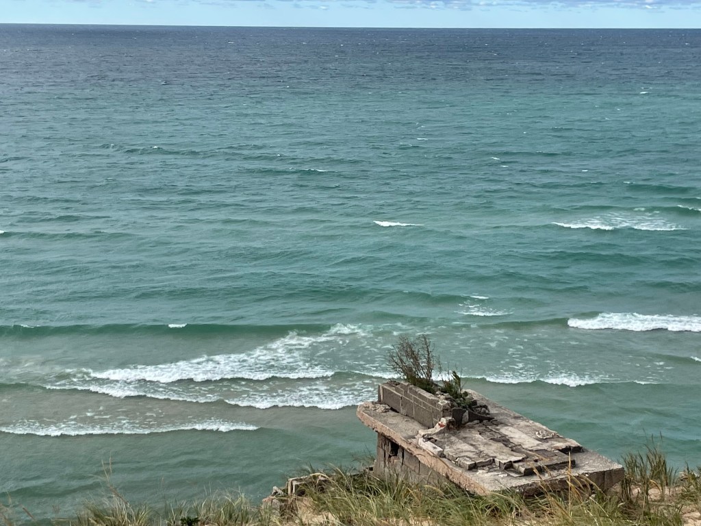 A concrete structure on a grassy hillside overlooking a calm sea with gentle waves under a cloudy sky.