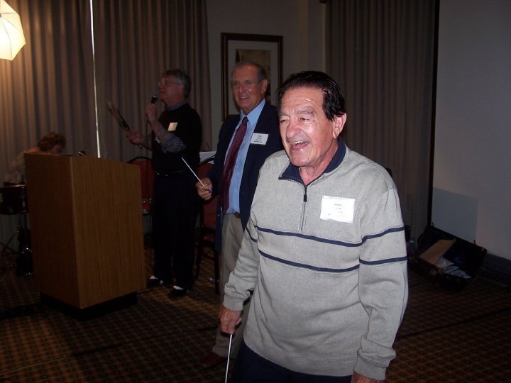 A happy older man in a gray sweater walks in front of a podium, smiling at an event. Two other men are visible in the background, one speaking into a microphone and the other watching.
