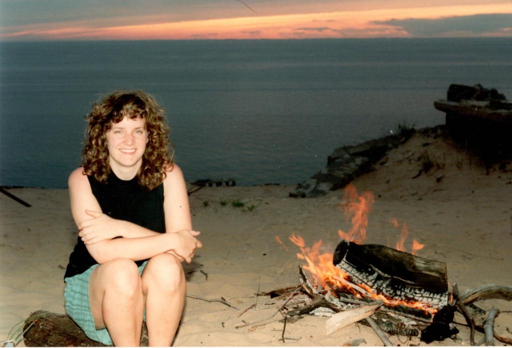 Smiling woman sitting by a campfire on the beach at sunset, with the ocean in the background.