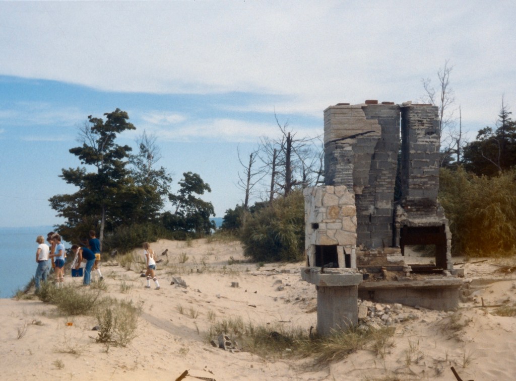 A group of people standing near the ruins of a stone fireplace on a sandy beach, with trees and a body of water in the background.