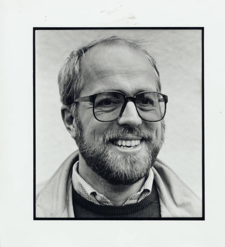 Black and white portrait of a smiling man with glasses and a beard, wearing a collared shirt and sweater.