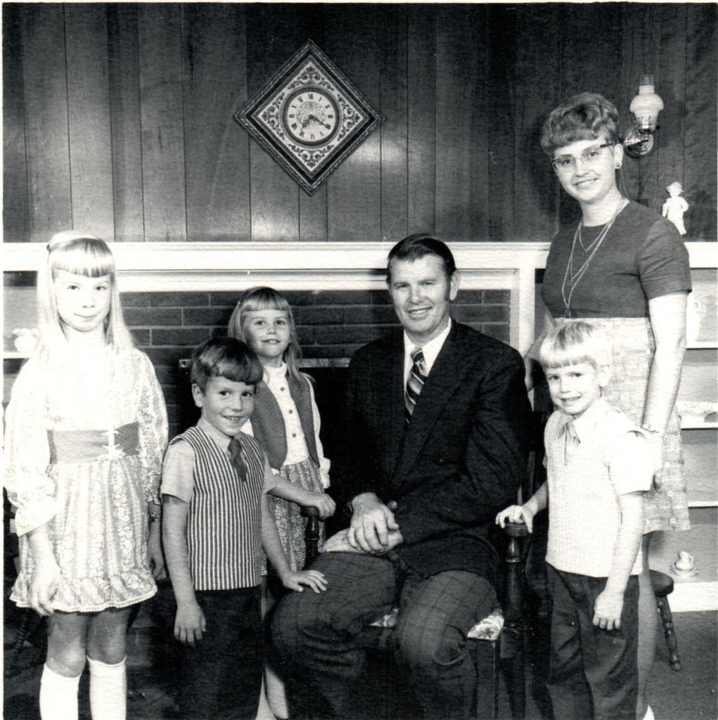 A black and white photo of a family group standing and sitting together in a living room, featuring four children and two adults. The children, dressed in retro clothing, are positioned around a man in a suit seated on a chair, while a woman in a vintage dress stands beside him.