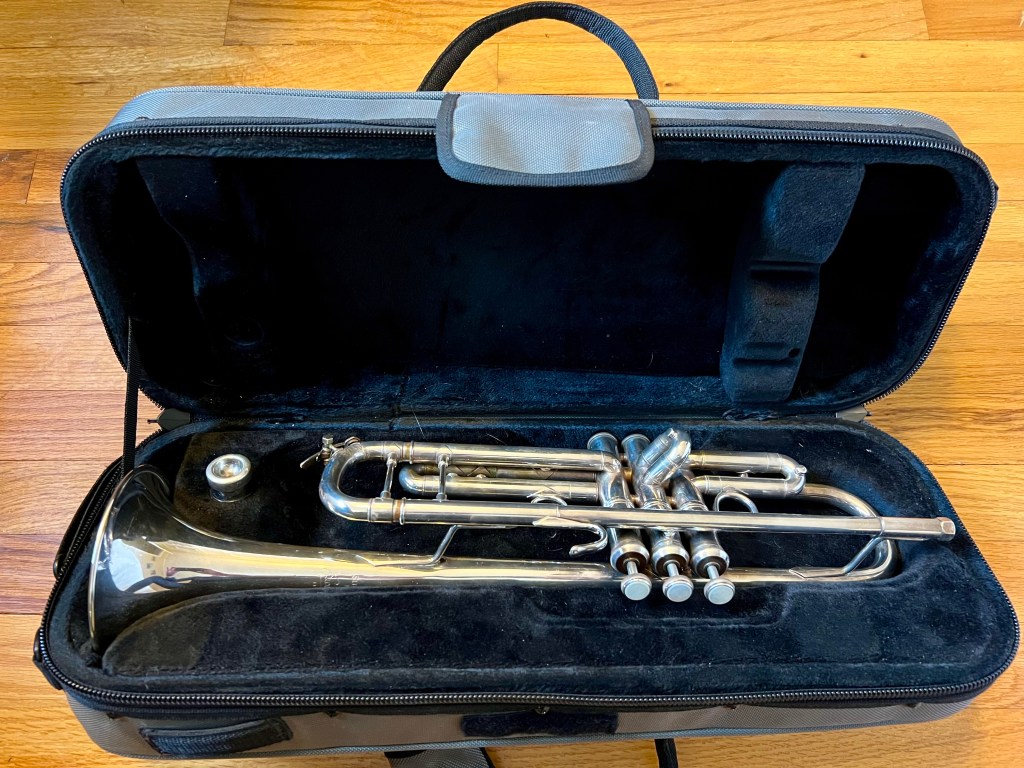A silver trumpet resting in a black, cushioned case with a zippered closure.